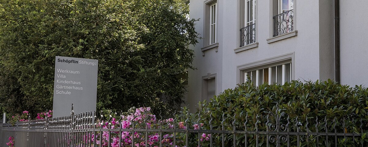[Translate to English:] Ein Gebäude mit hellgrauen Wänden und mehreren Fenstern wird teilweise von Bäumen und Büschen verdeckt. An einem Metallzaun hängt ein Schild mit der Aufschrift "Schöpflin Stiftung", "Schöpflin Schule" und aktuellen Neuigkeiten zu den verschiedenen Einrichtungen.