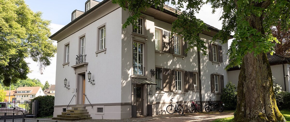 Ein zweistöckiges weißes Gebäude mit großen Fenstern, einer Treppe, die zum Eingang führt, und mehreren Fahrrädern, die draußen geparkt sind, umgeben von Bäumen und Grünflächen.