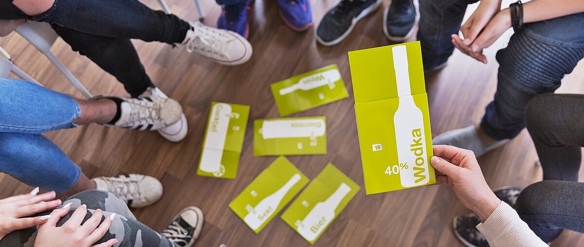 Eine Gruppe von Personen sitzt im Kreis und hält gelbe Karten in der Hand, auf denen eine Flasche abgebildet ist und das Wort Wodka neben 40% steht.